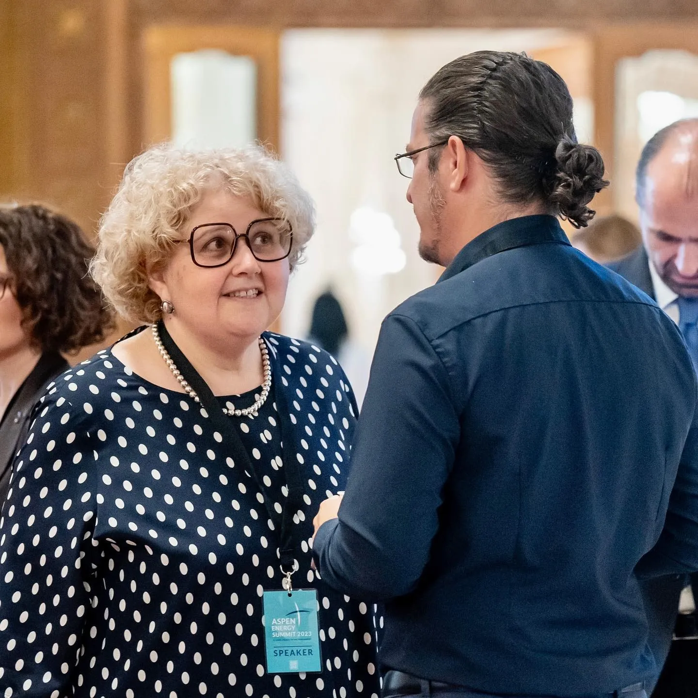 Woman with curly blonde hair and glasses in a polka-dot top with an Aspen Energy Summit 2023 speaker badge talks with a man in a dark blue shirt.