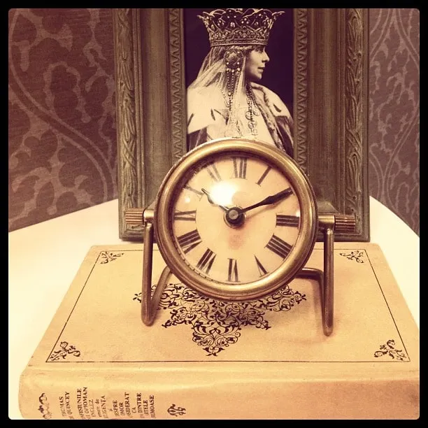 A brass mantel clock resting on an old book, in front of a framed portrait of a woman wearing a crown.