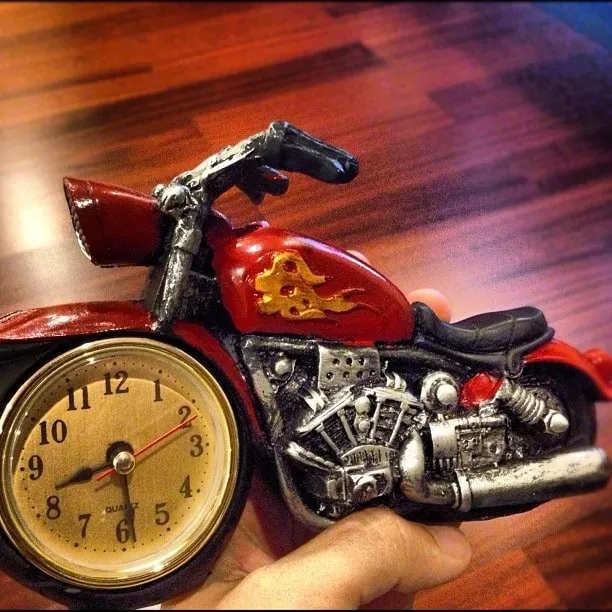 A hand holds a red motorcycle-shaped desk clock on a wooden countertop.