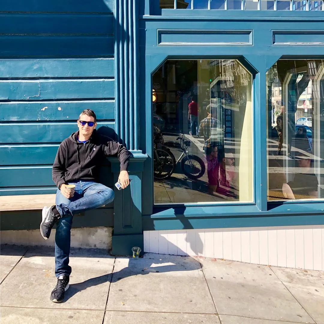 Man in sunglasses and a black hoodie leans against a teal storefront holding a coffee cup on a sunny sidewalk