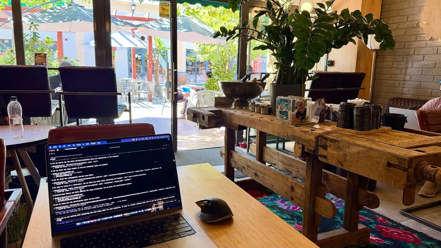 A laptop and mouse sit on a wooden table in a bright café filled with rustic furniture and plants.