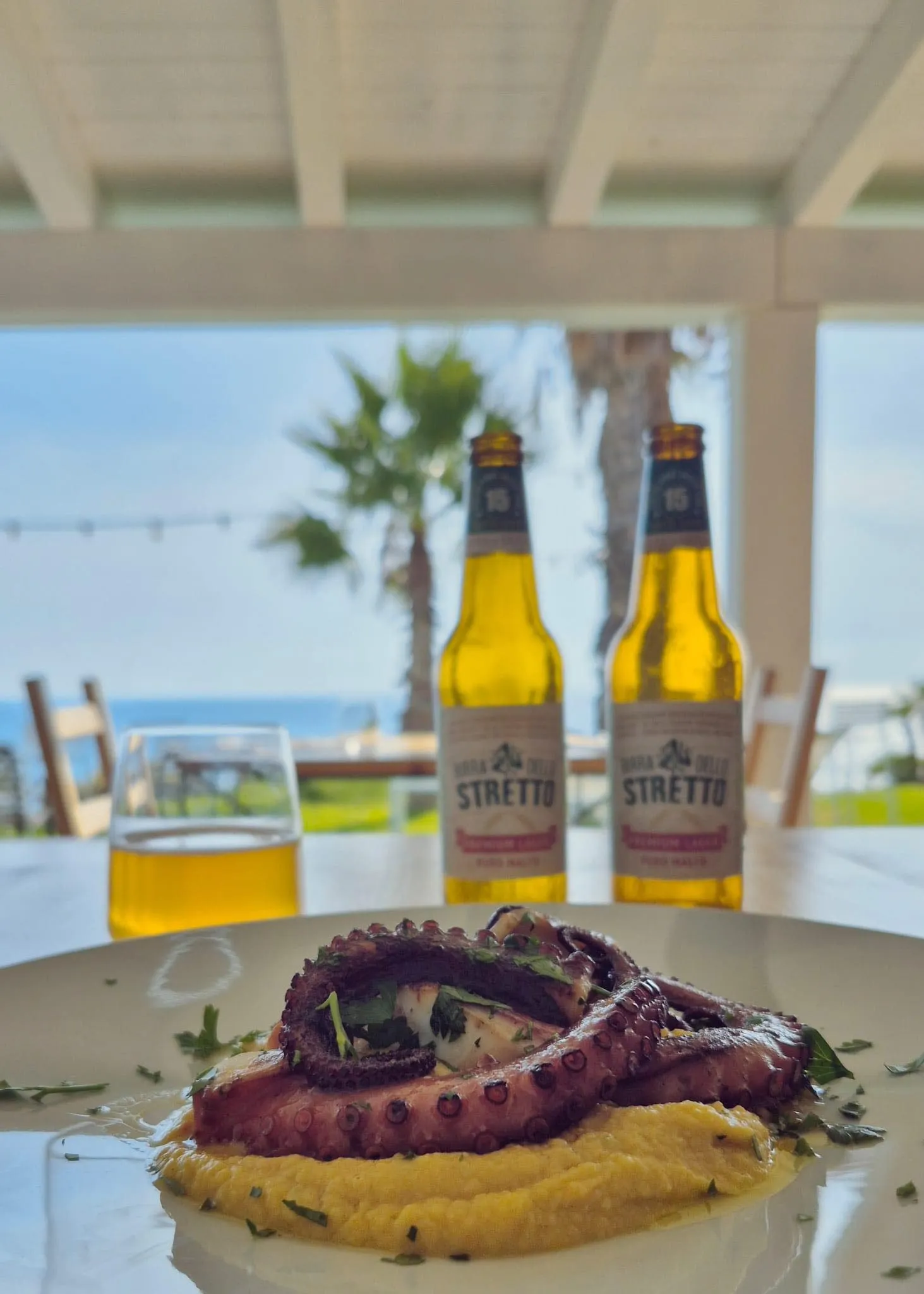 A plate of grilled octopus and two beer bottles sit on a seaside restaurant table.