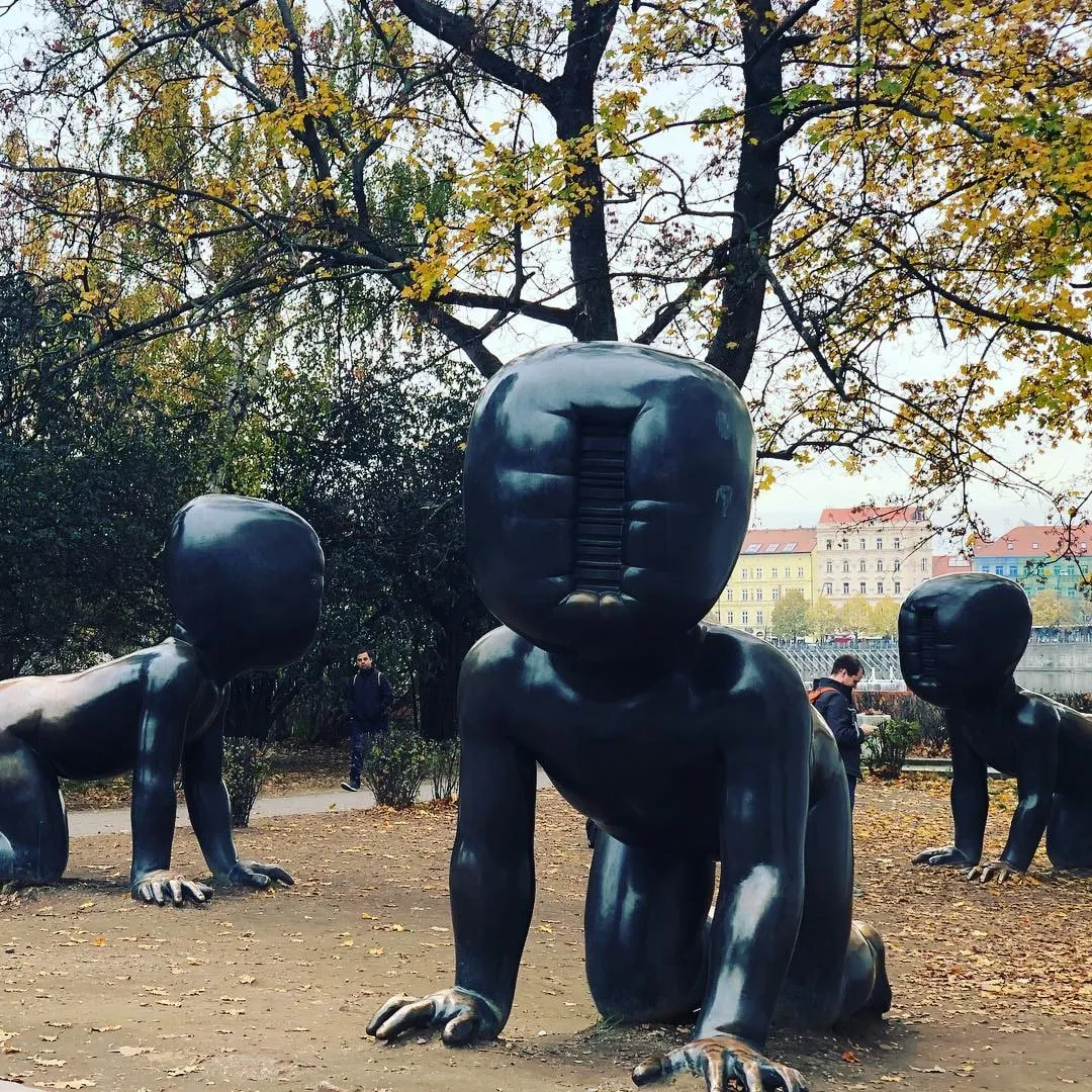 Three giant black crawling baby sculptures with faceless heads in an autumn park among fallen leaves and trees
