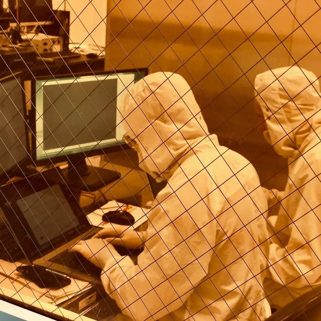 Two people in white cleanroom suits work at computers and a laptop behind a diamond-pattern wire mesh