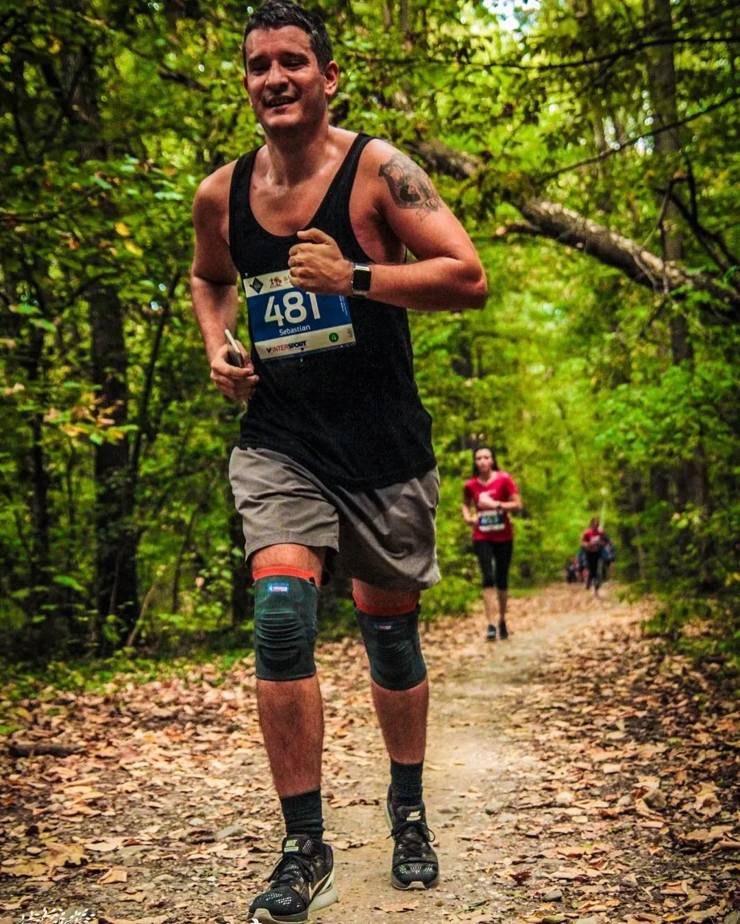 A man in a black tank top, race bib 481, and knee braces runs along a leaf-covered forest trail