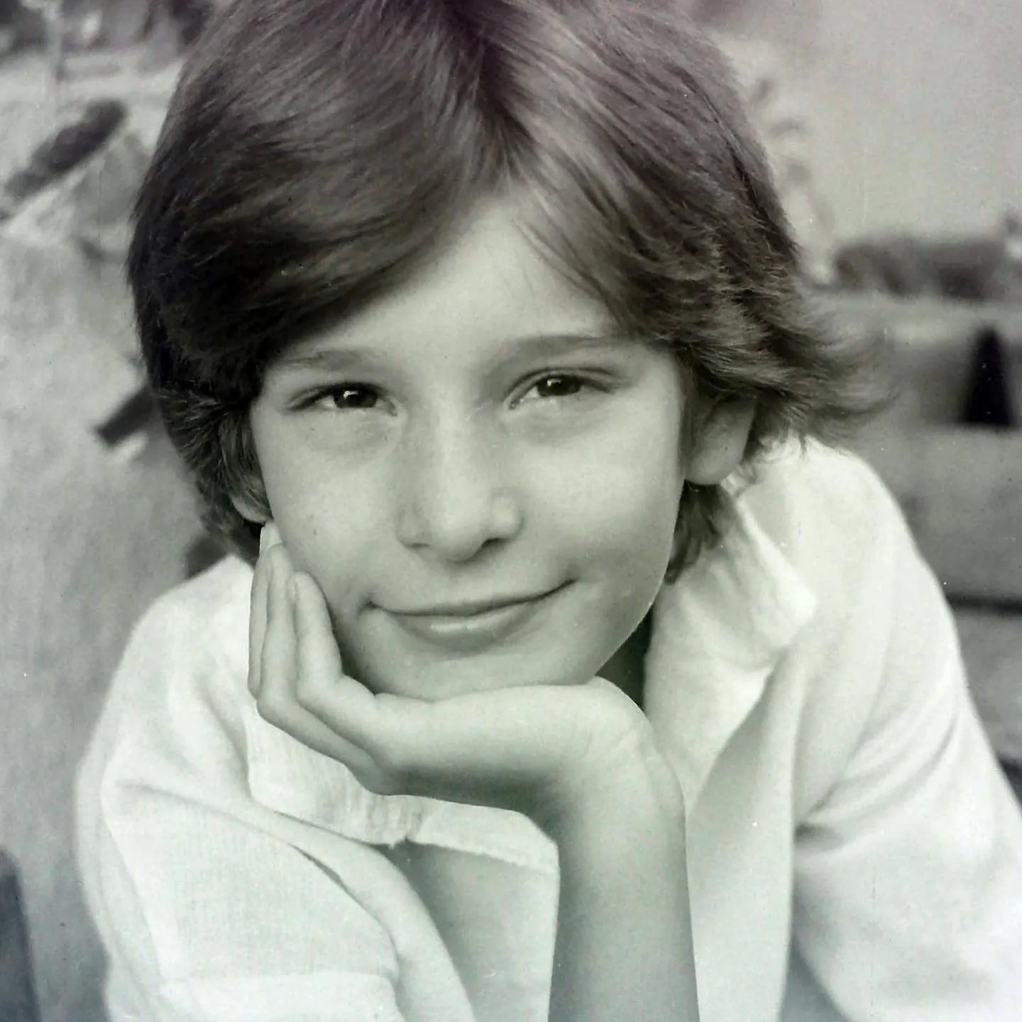 A black-and-white childhood portrait shows a smiling boy resting his face on his hand.