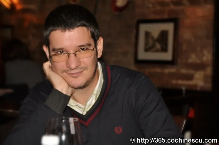 A man with glasses smiles, resting his head in his hand at a table.