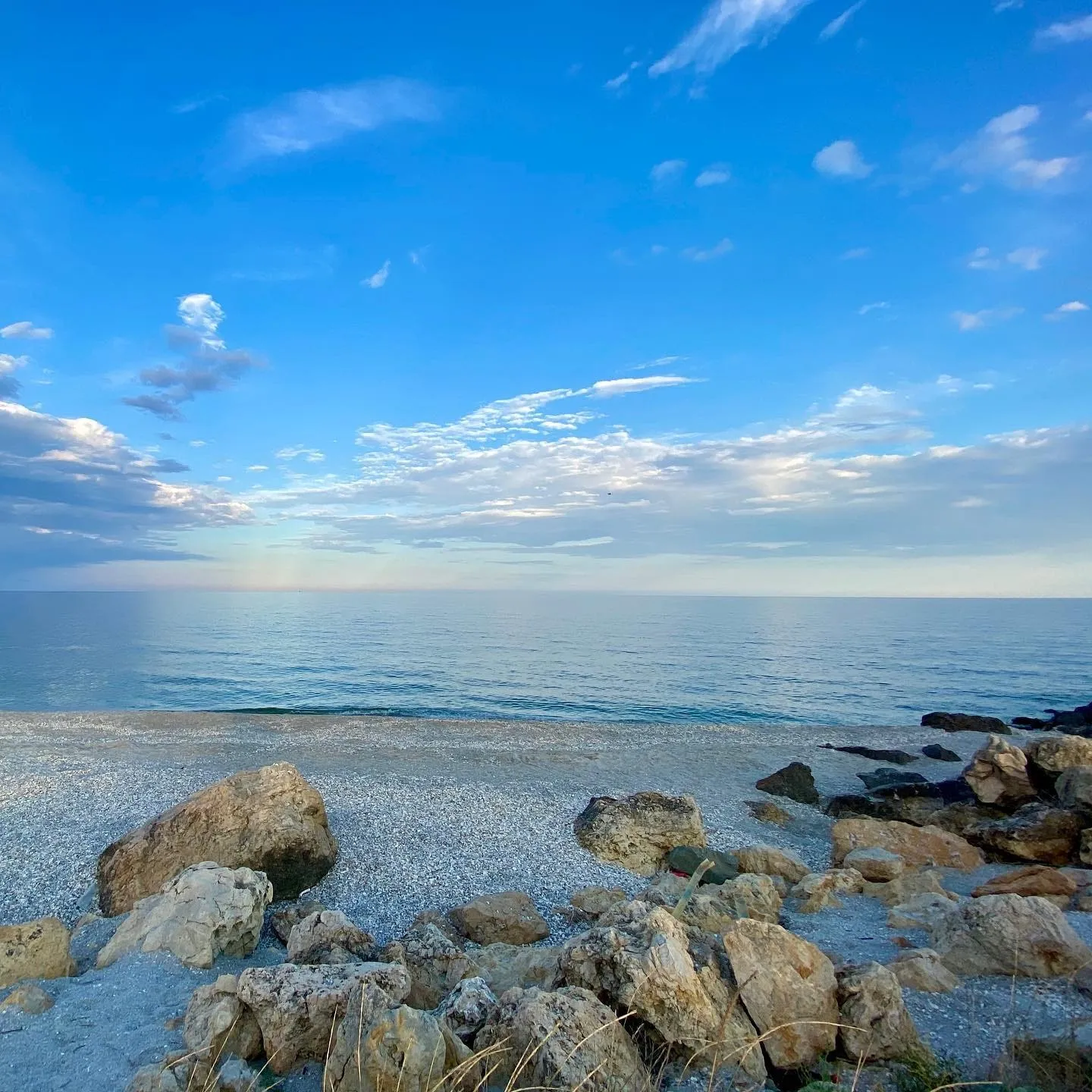 A quiet rocky beach meets a calm blue sea beneath a wide sky with scattered clouds.