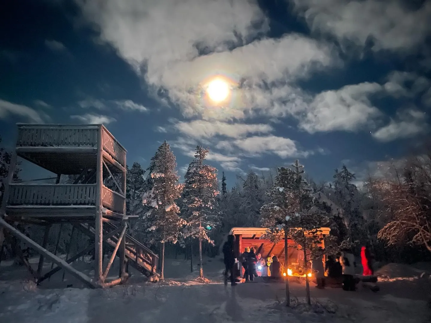 A bright moon shines through clouds above a snowy forest gathering lit by a warm fire.