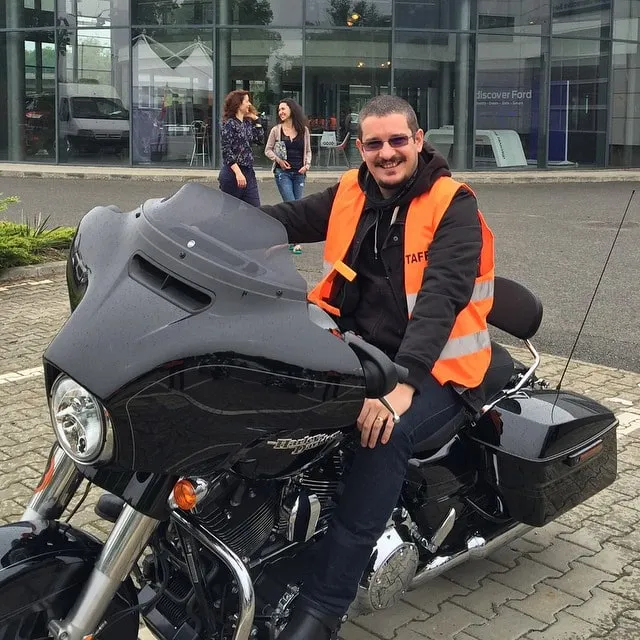 Smiling man on a black Harley-Davidson motorcycle, wearing an orange reflective vest and sunglasses, in front of a modern building.