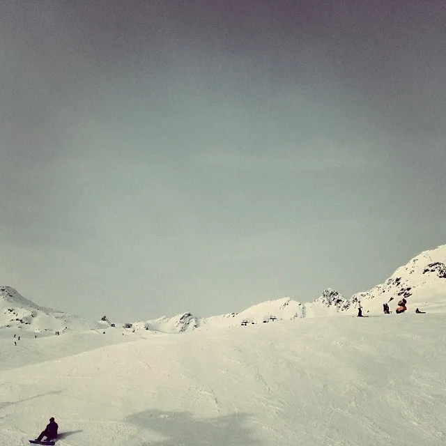 A wide snowy ski slope stretches toward mountain peaks under a pale overcast sky, with a few distant skiers.