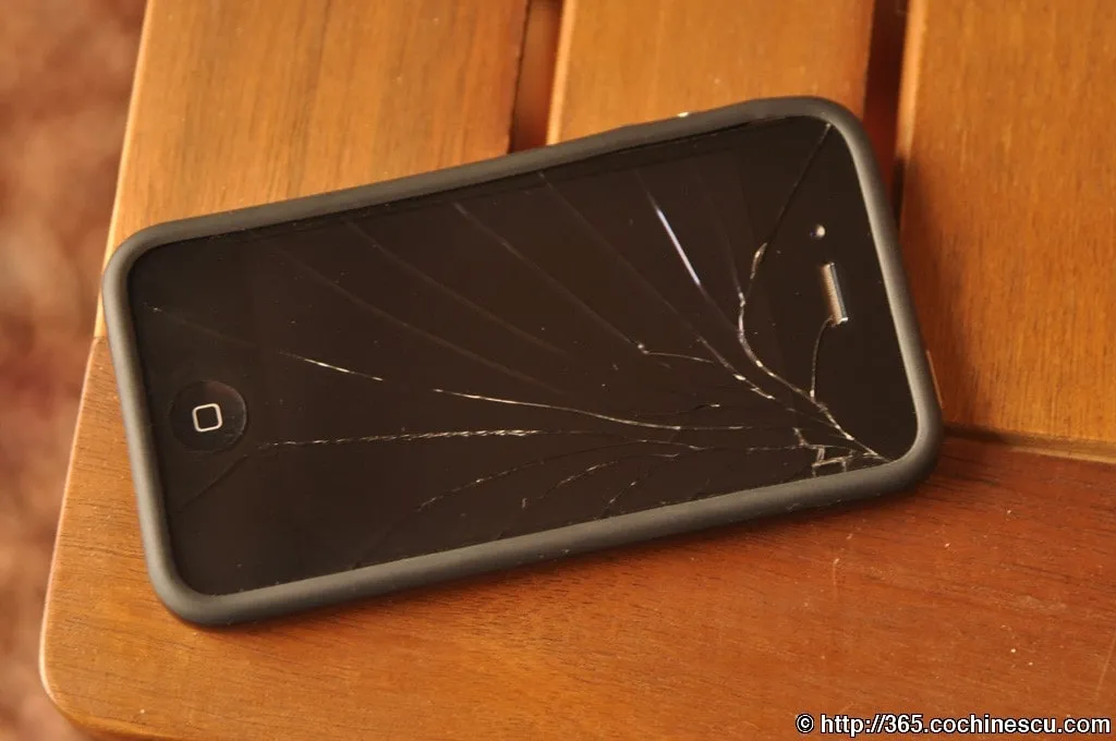 An iPhone 4 with a broken screen is sitting on a wooden table.