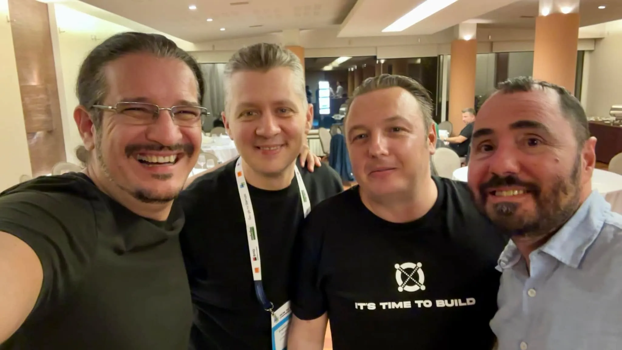 Four men smile for a close-up selfie in a banquet hall, one wearing a conference lanyard and another an IT’S TIME TO BUILD shirt.