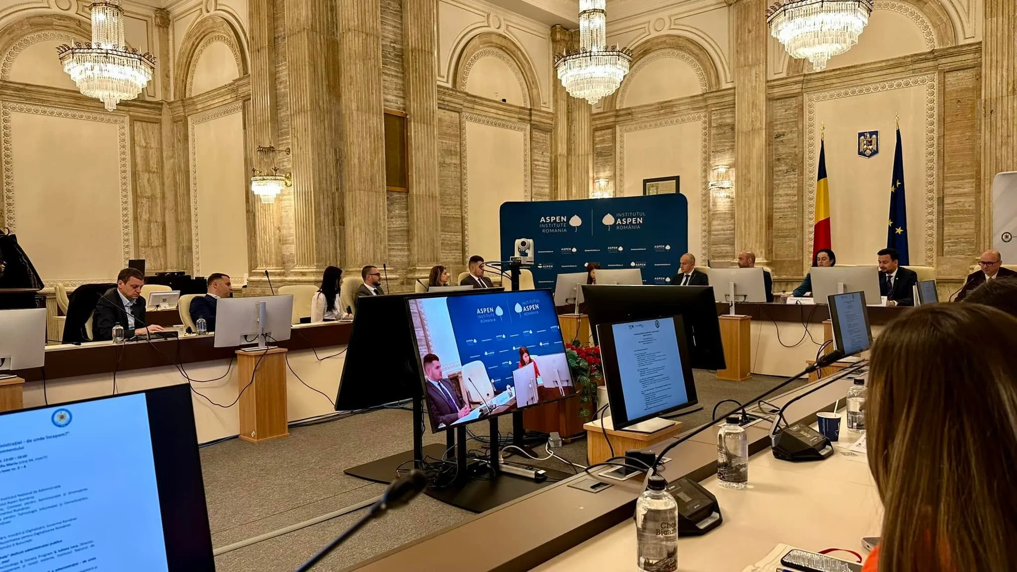 Participants seated at a U-shaped conference table in an ornate hall with Aspen Institute Romania banners and Romanian and EU flags.