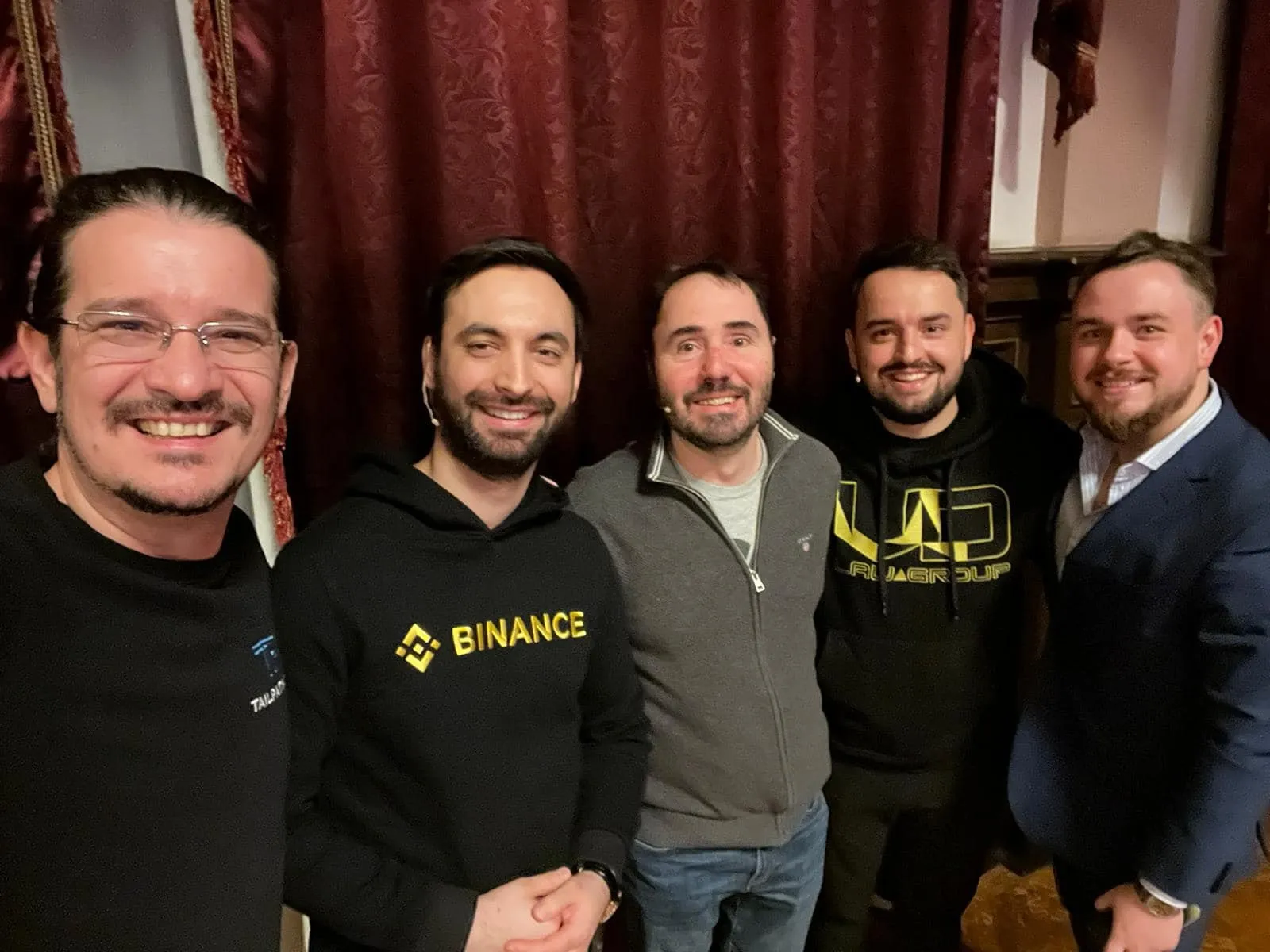 Five men smile for a group photo indoors, two wearing Binance and NU Group hoodies against red curtains.