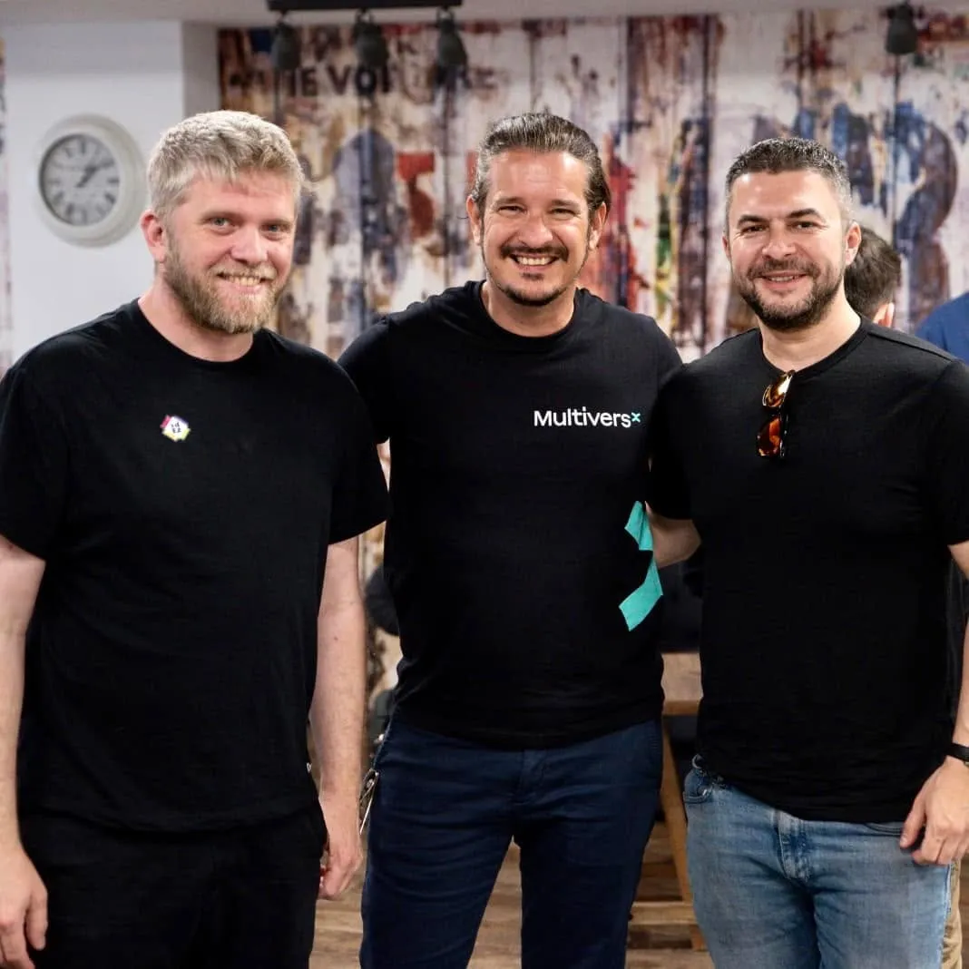 Three smiling men in black t-shirts pose together indoors, the middle one wearing a MultiversX logo shirt.