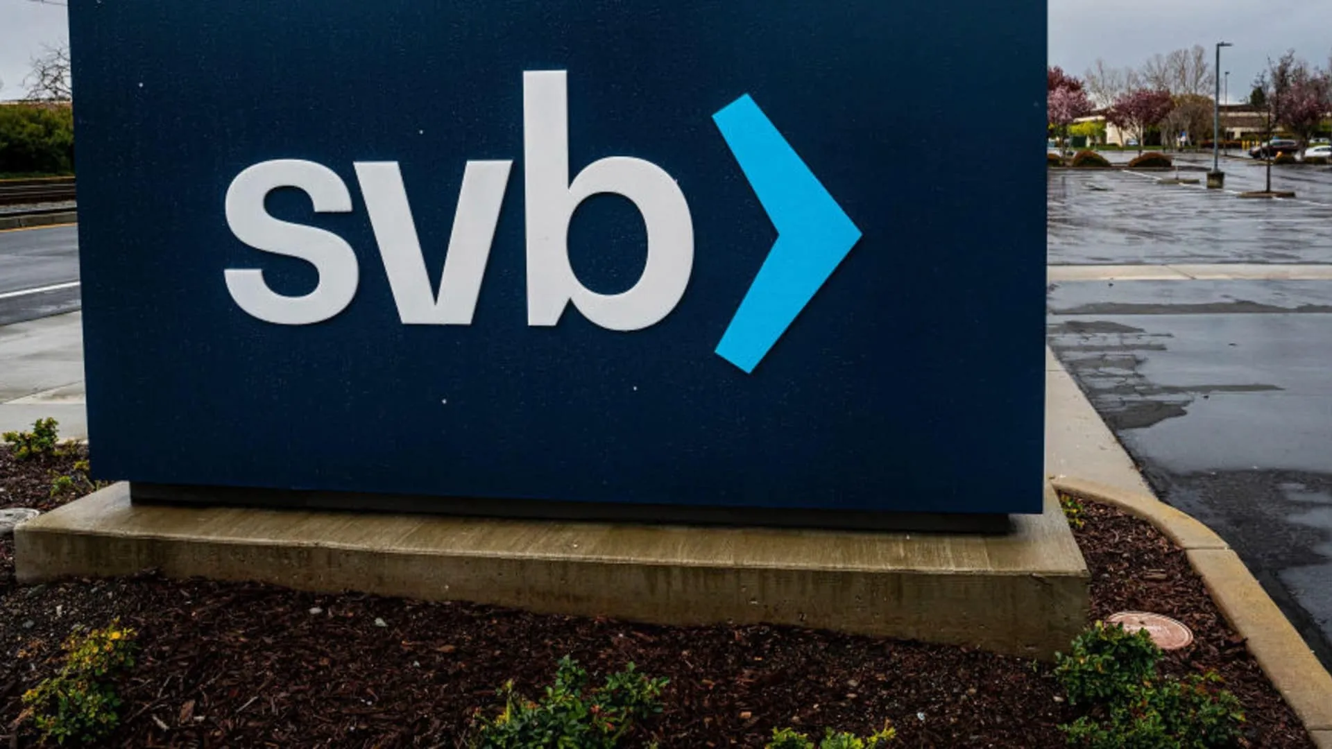 Close-up of a large blue outdoor Silicon Valley Bank sign beside a wet parking lot under an overcast sky.