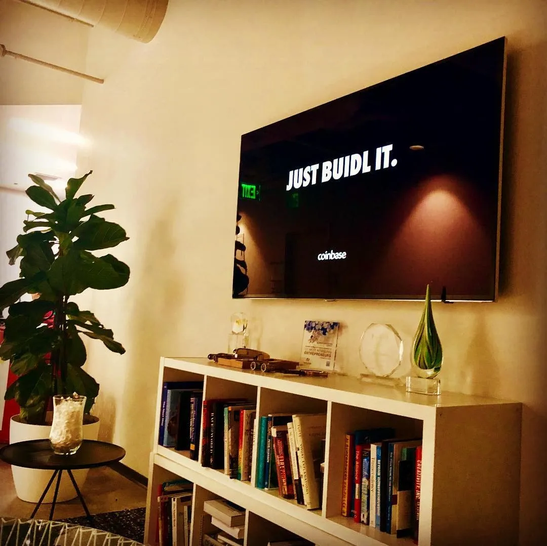 Wall-mounted TV showing Just BUIDL IT and the Coinbase logo above a white bookshelf with awards and plants