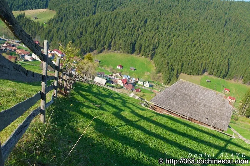 A mountain village seen from a green hillside, next to a fence and a wooden barn.
