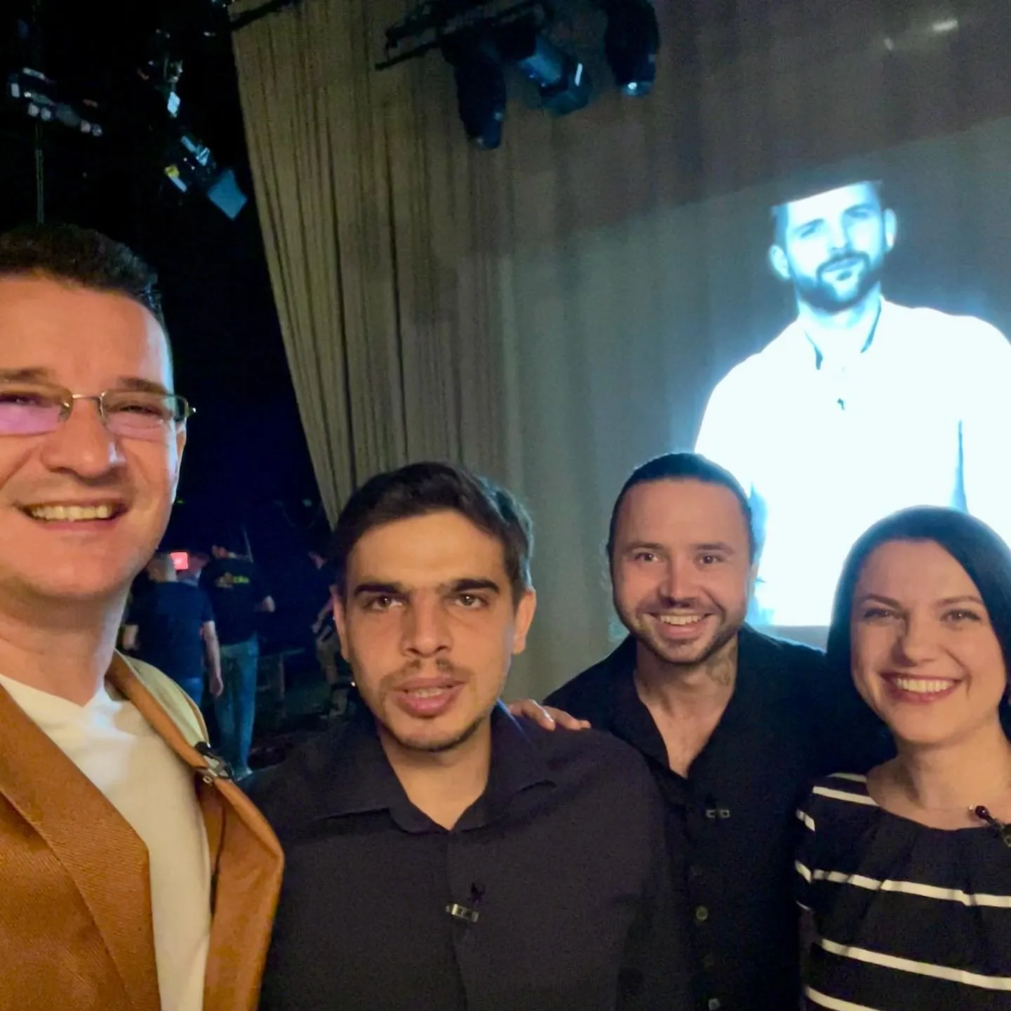 Four people pose for a selfie backstage in front of a large illuminated screen.