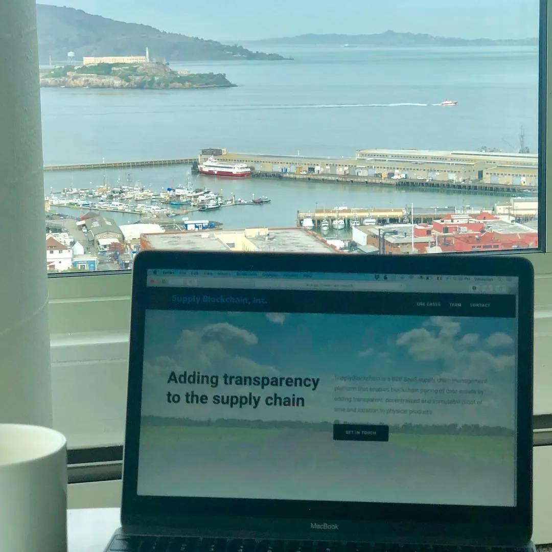 Open MacBook showing the Supply Blockchain website on a windowsill overlooking San Francisco Bay, a marina, and Alcatraz