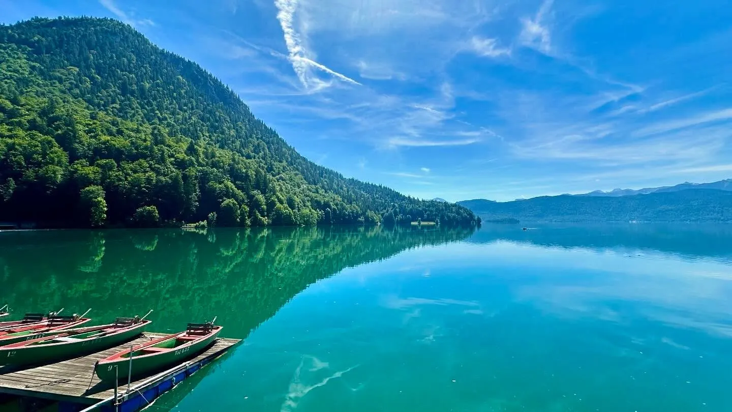 Wooden rowboats rest on a dock beside a calm turquoise lake with forested hills under a blue sky.