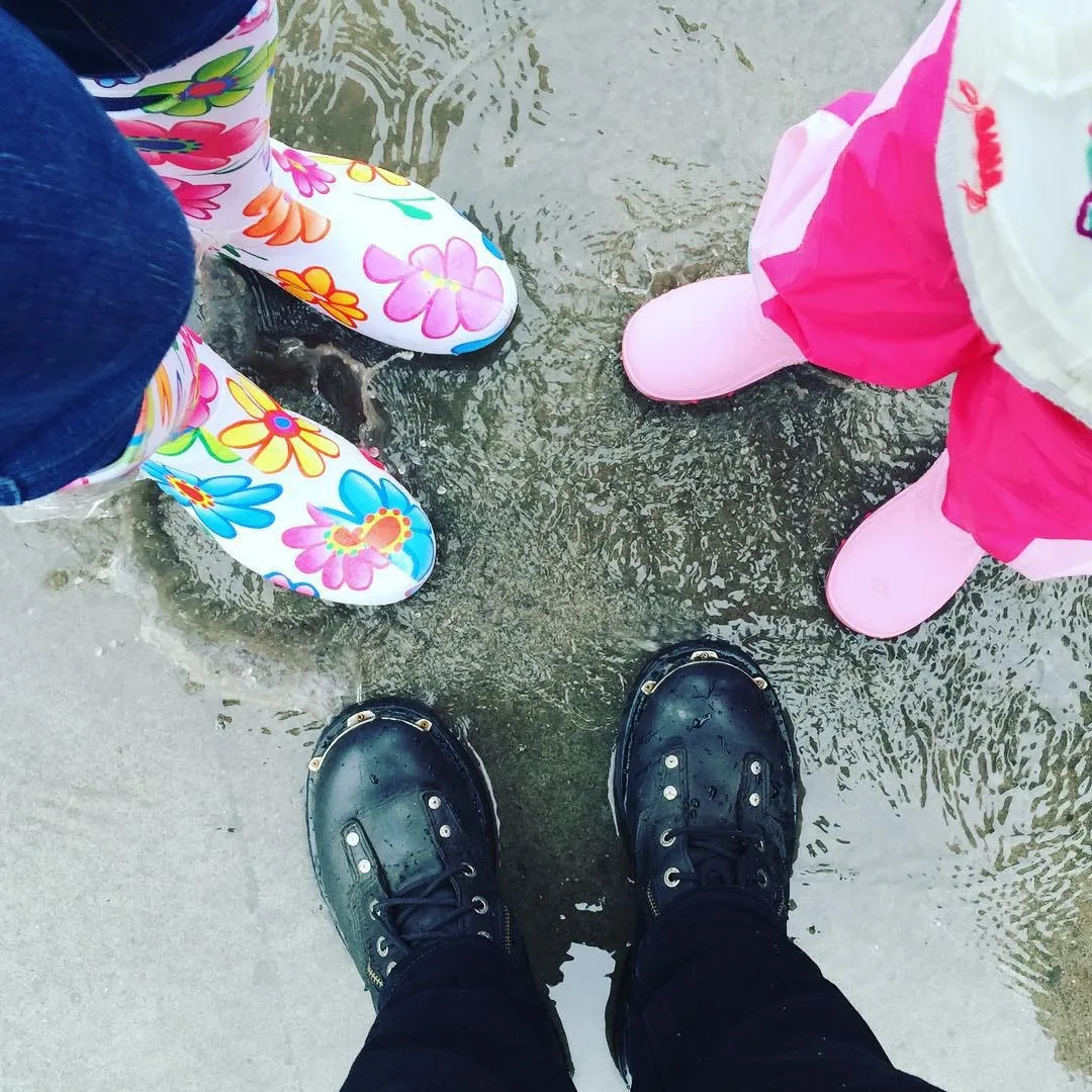 Three pairs of feet in colorful rain boots and black boots stand in a puddle on the asphalt.