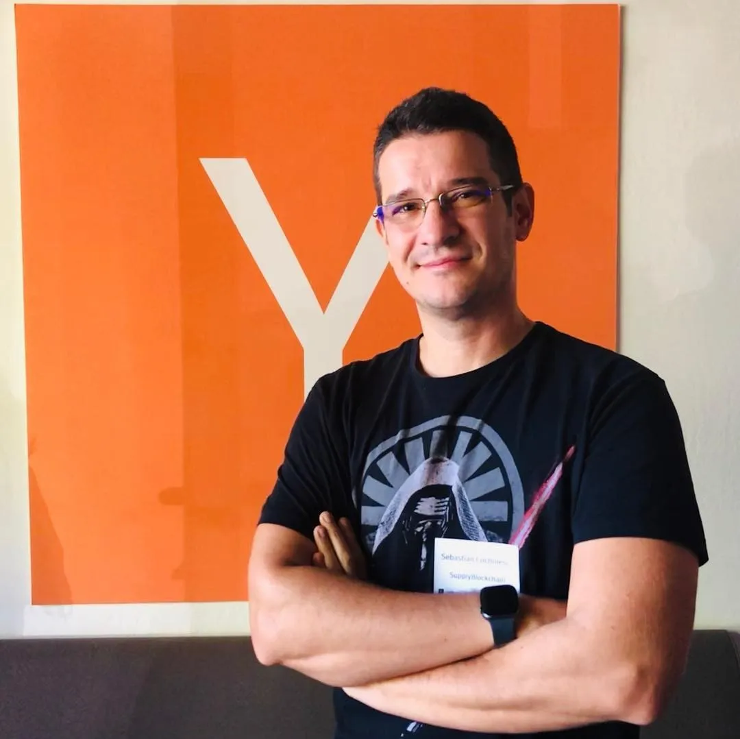 Man in a black graphic t-shirt and name badge standing arms crossed in front of a large orange Y Combinator logo