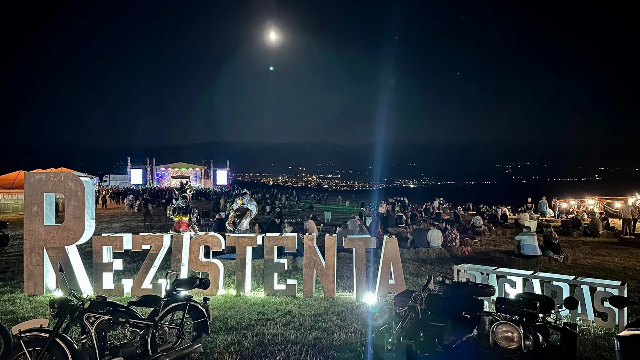 Illuminated Rezistenta and Brigadas letters at a nighttime outdoor festival with motorcycles, a crowd, and a moonlit stage.