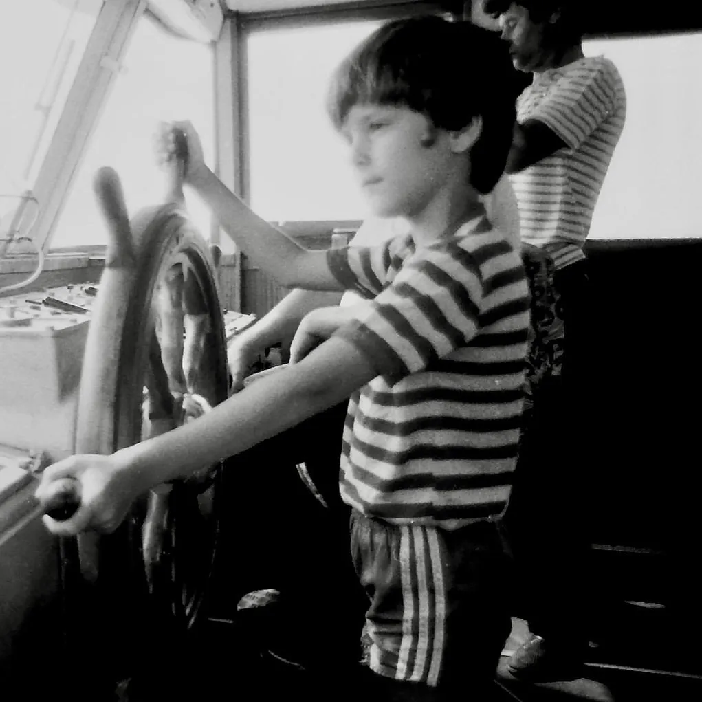A young boy stands at a ship’s wheel in a black-and-white childhood photograph.
