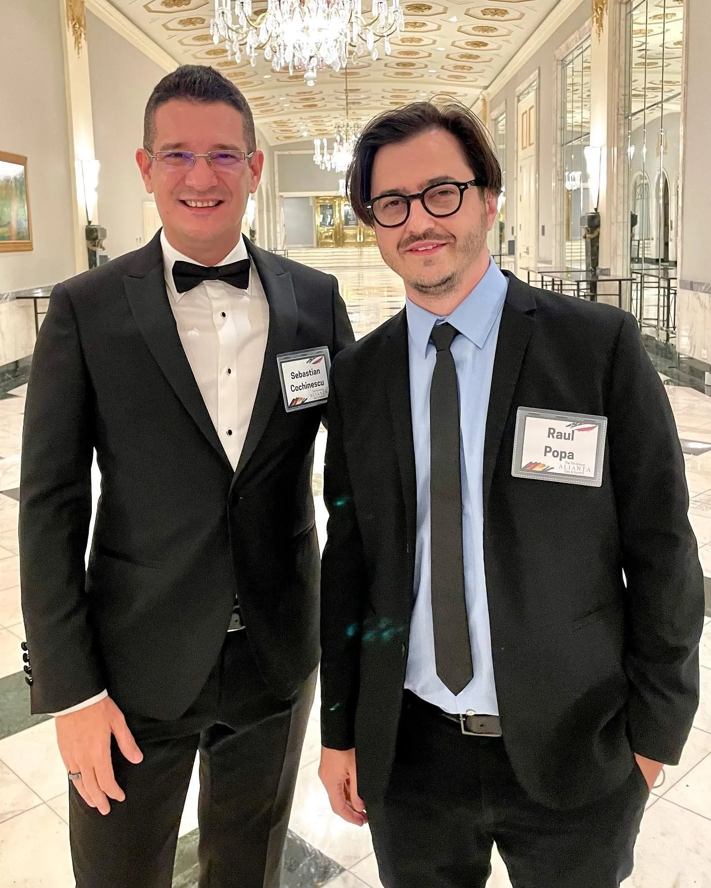Sebastian Cochinescu and Raul Popa stand together in formal attire inside an elegant chandelier-lit hall.