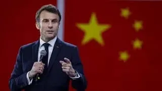Emmanuel Macron in a dark suit speaks into a microphone in front of a red Chinese flag with yellow stars.