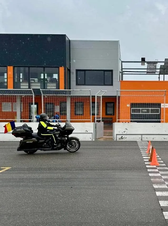 A motorcyclist on a black touring bike with a Romanian flag rides past orange cones near a checkered finish line.