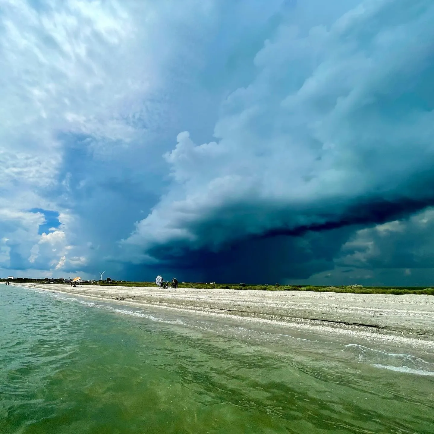 Massive dark storm clouds approach a beach and green-blue coastal water.