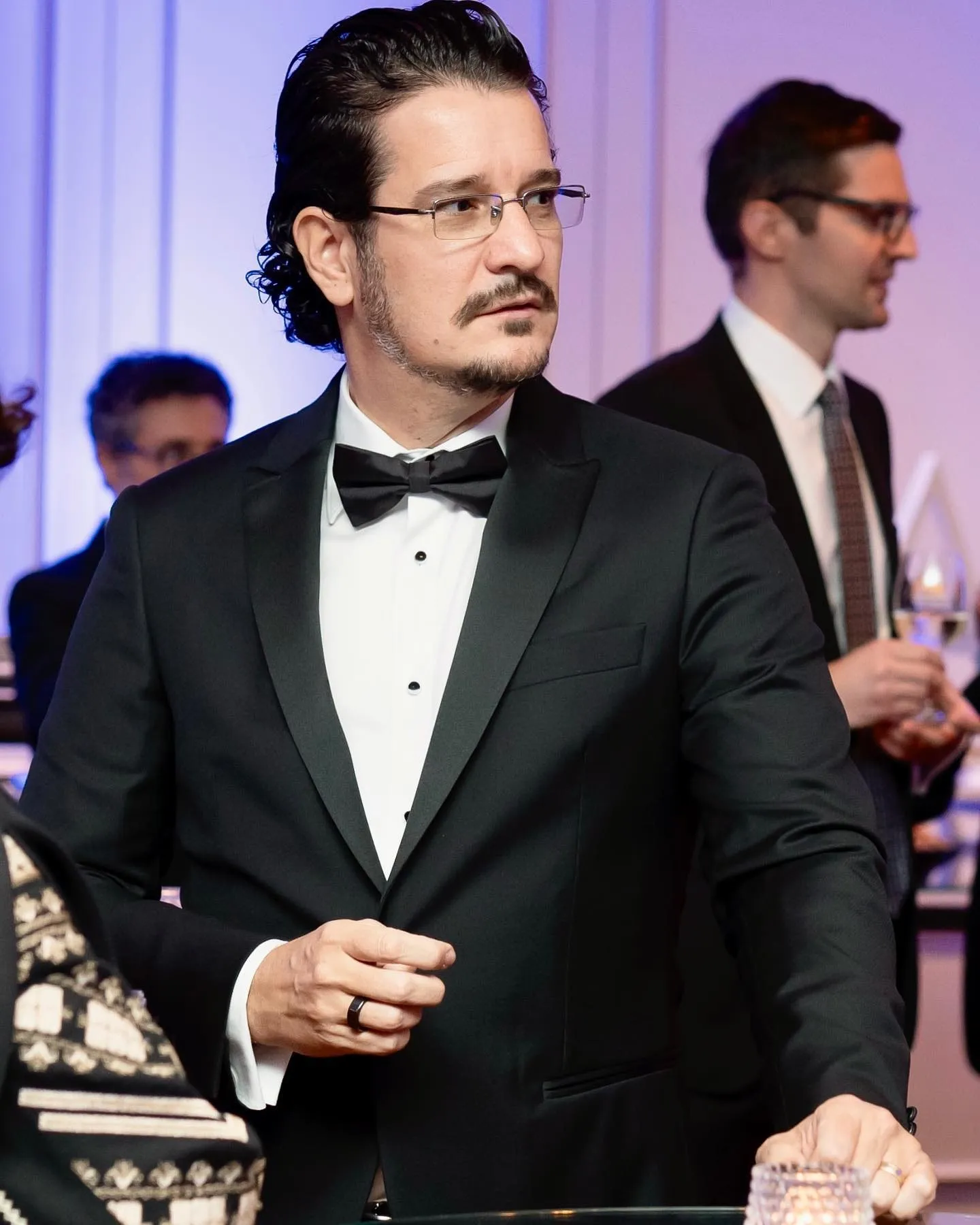 A man in a black tuxedo and bow tie stands at a formal reception among other guests.