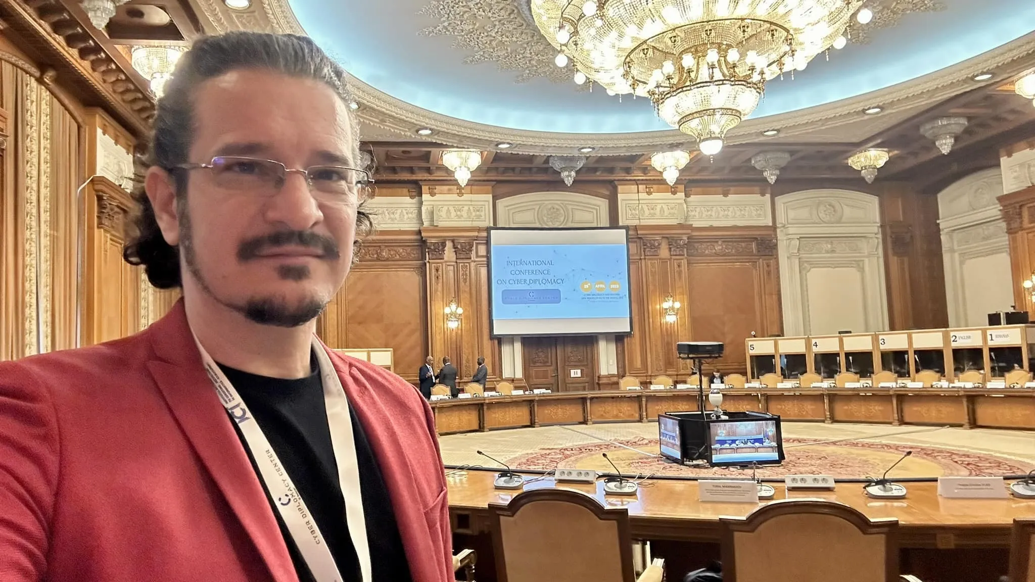 A man in a red blazer and conference badge takes a selfie in an ornate hall with a cyber diplomacy presentation screen.
