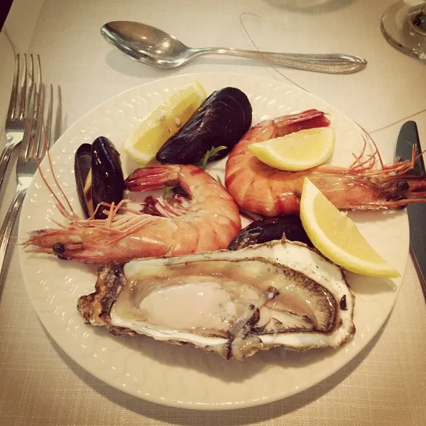 White plate with shrimp, mussels, an open oyster and lemon wedges on a restaurant table