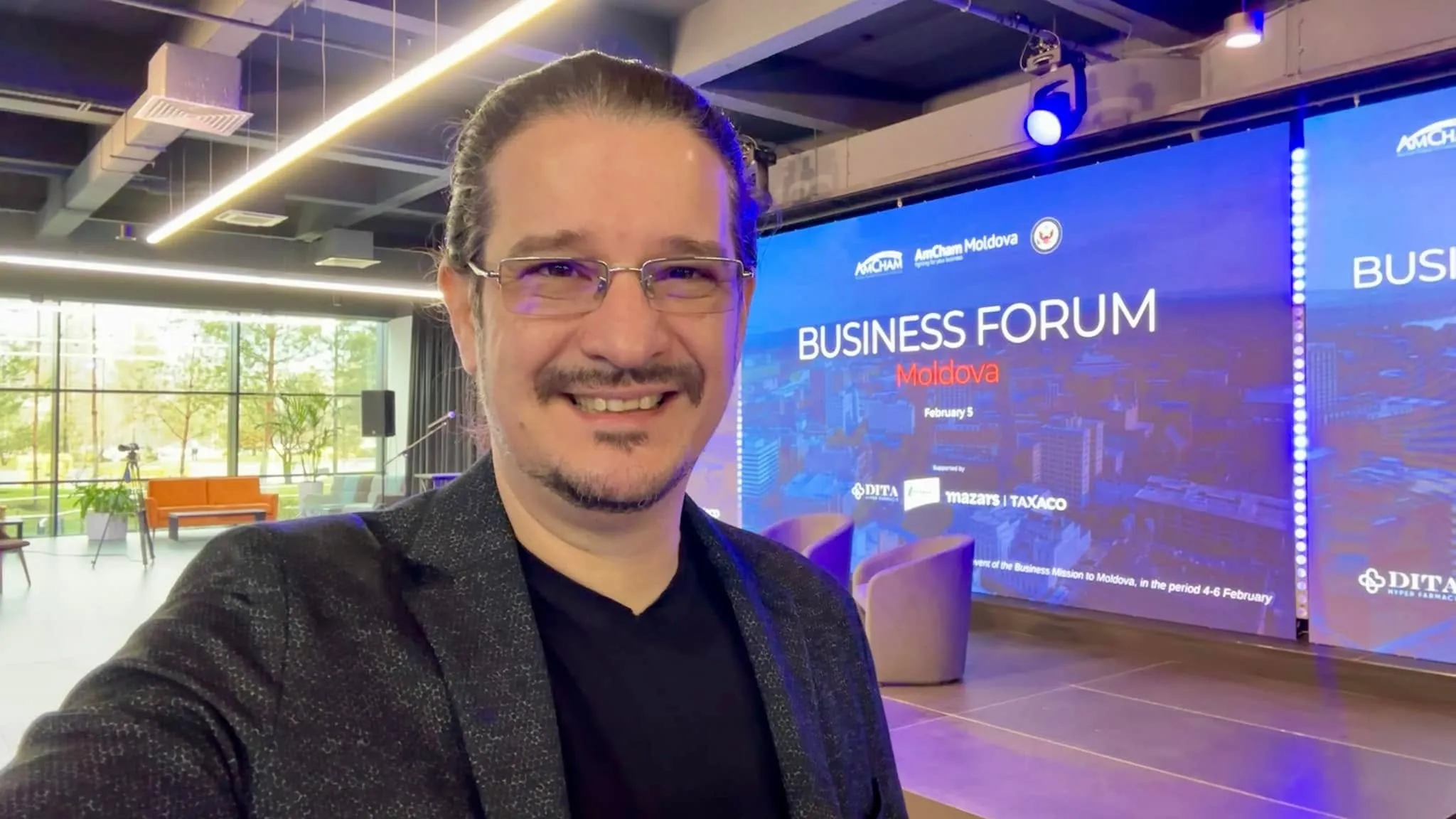 Sebastian Cochinescu smiles in a selfie at the Business Forum Moldova venue with the stage screen behind him.
