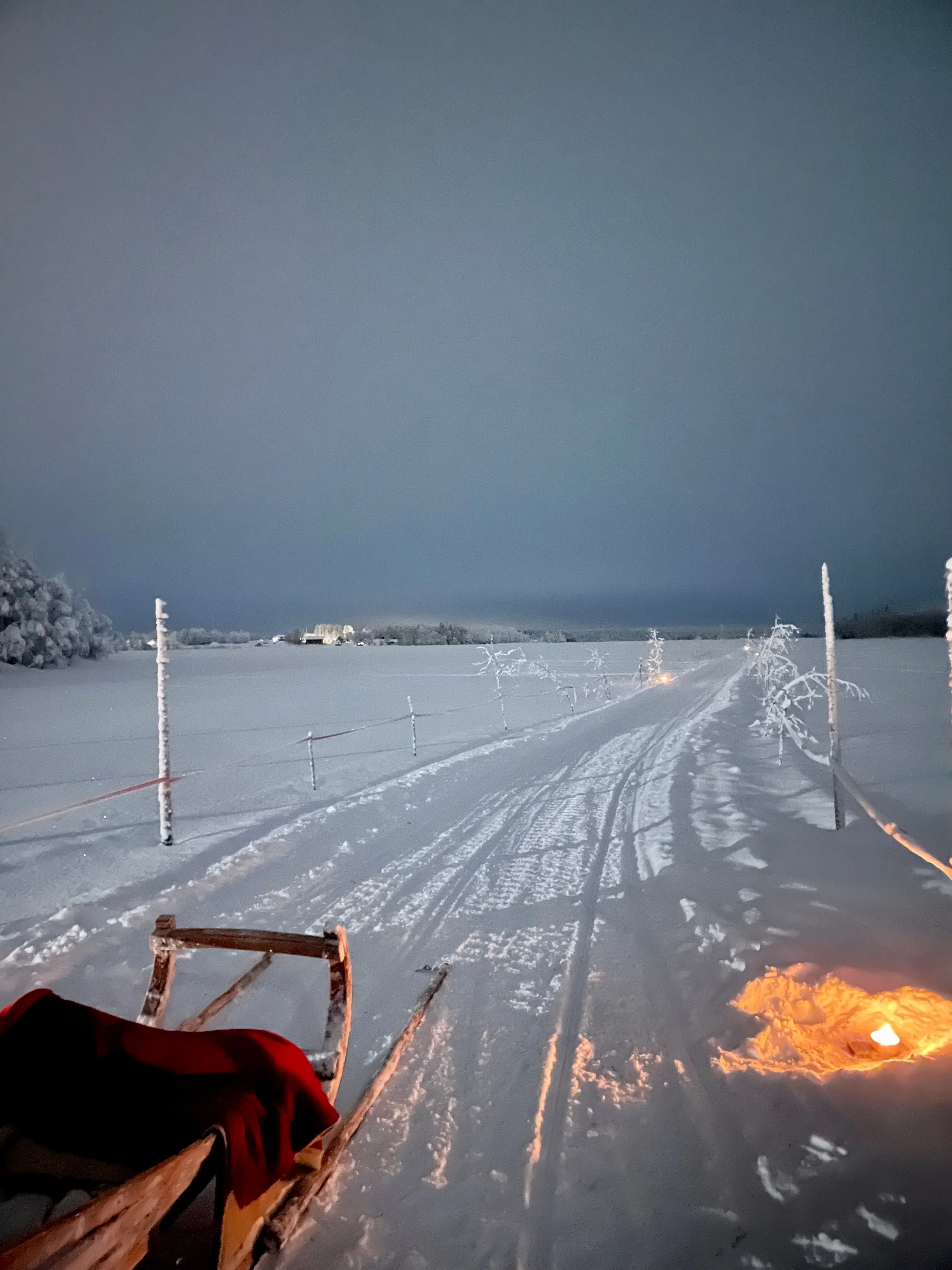 Sled trail across a snowy field at dusk, lined by small lights