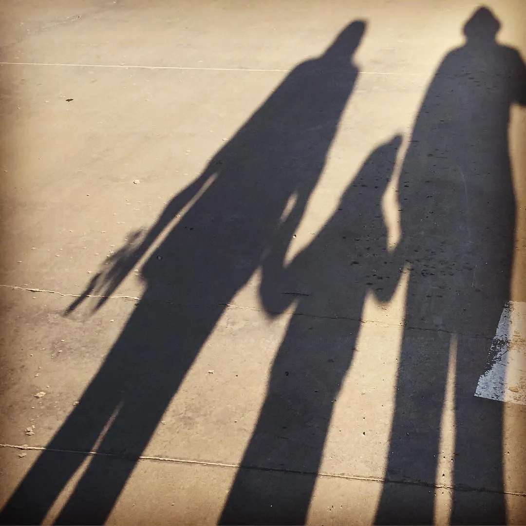 Three elongated shadows of people walking side by side on a sunlit concrete pavement.