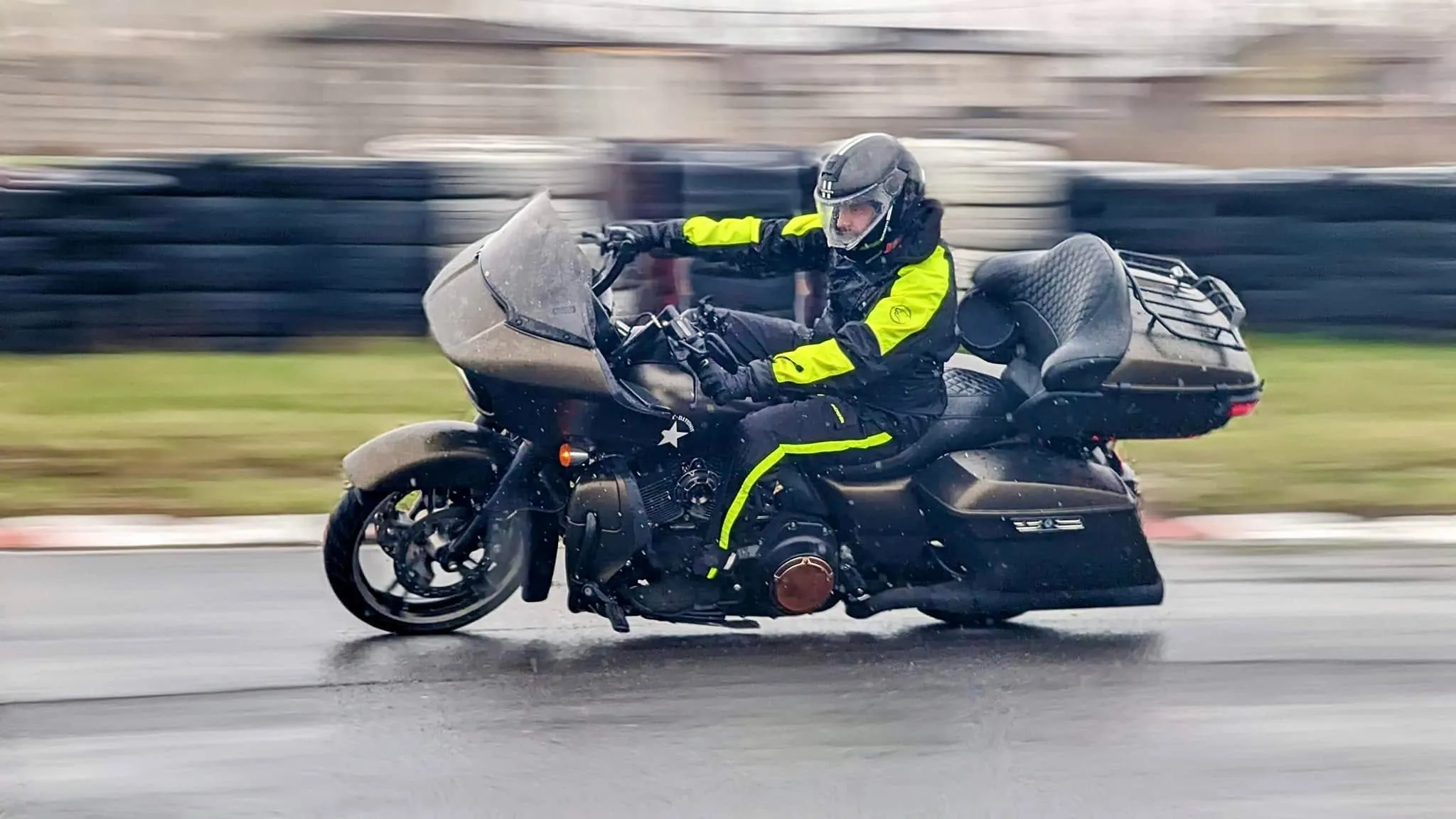 A motorcyclist in a black and neon yellow suit rides a touring bike on a wet track in the rain.