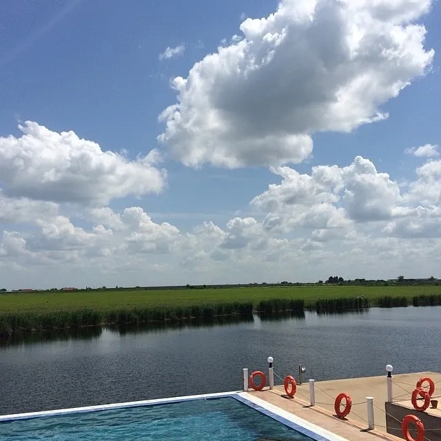 Piscină cu colaci de salvare portocalii, cu vedere spre un lac și câmpuri verzi sub un cer albastru cu nori albi.