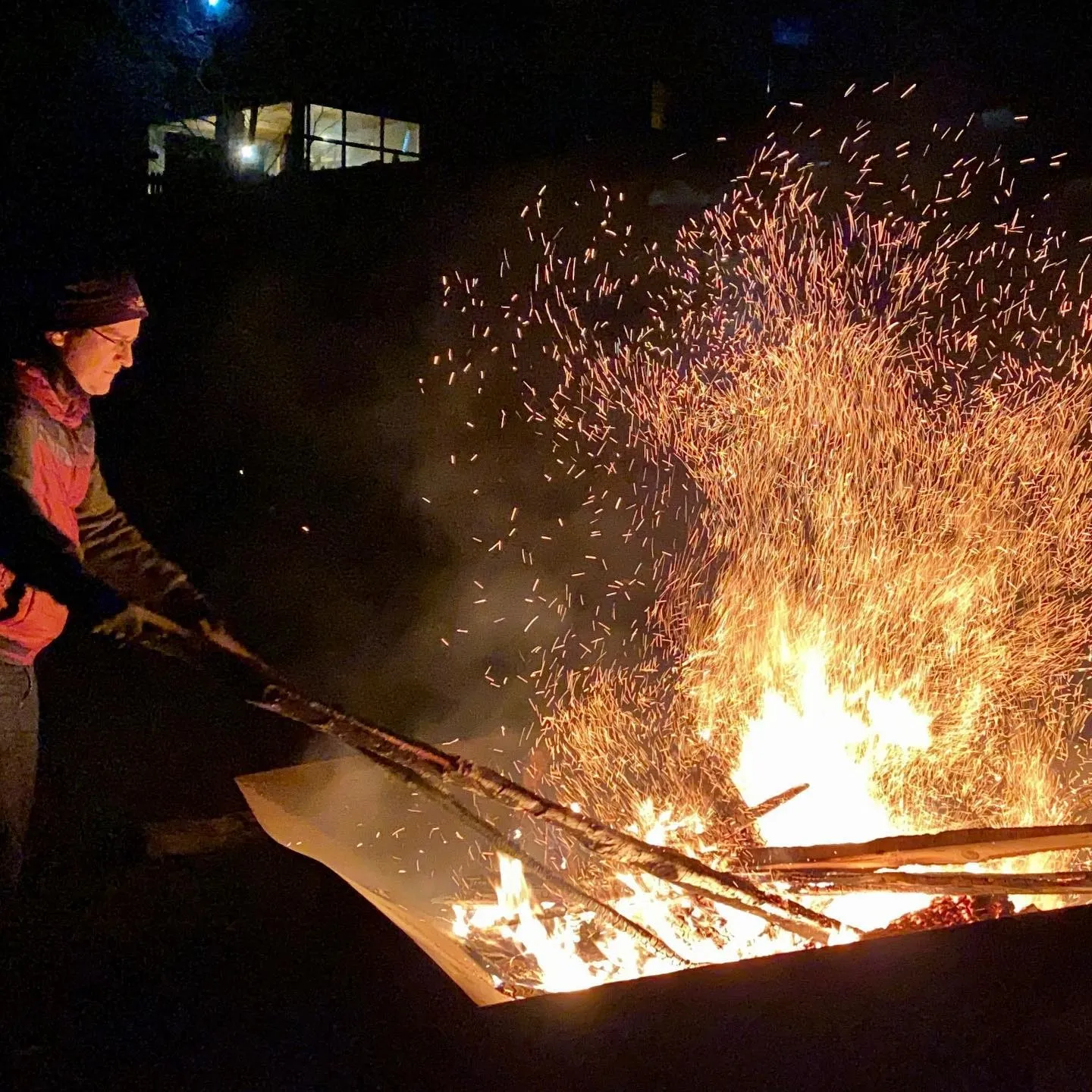 A person tends a large campfire as bright sparks rise into the night.