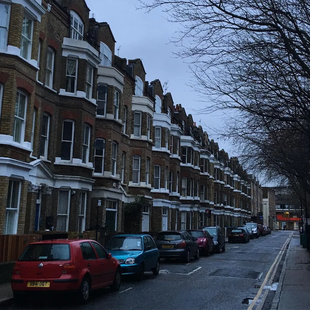 A row of brick terraced houses with bay windows and parked cars lining a wet London street.