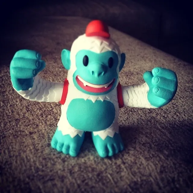 A teal and white Mailchimp Yeti Freddie figurine with raised arms standing on a brown fabric surface.