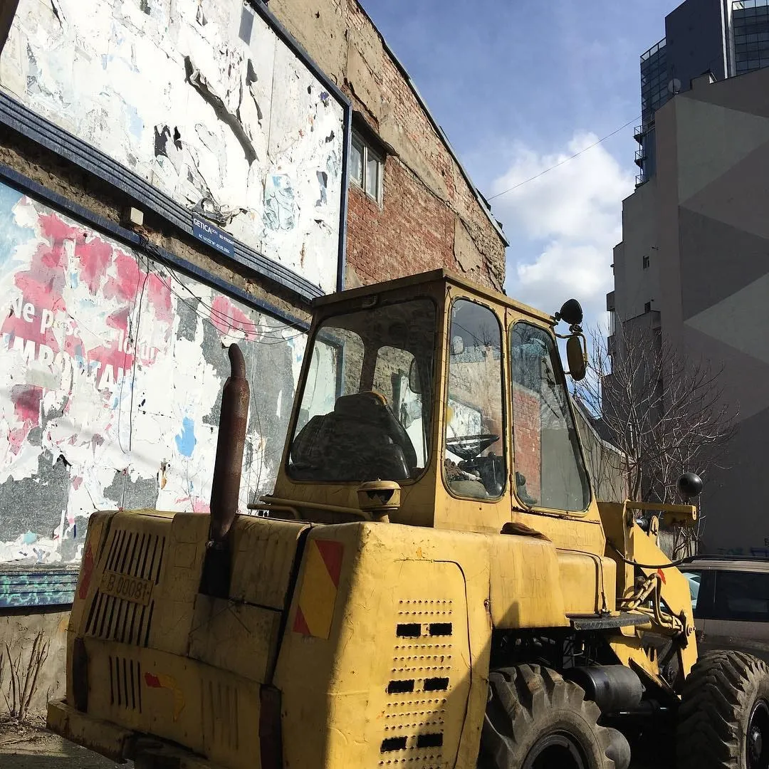 A weathered yellow vintage loader sits beside a peeling brick wall under a partly cloudy sky