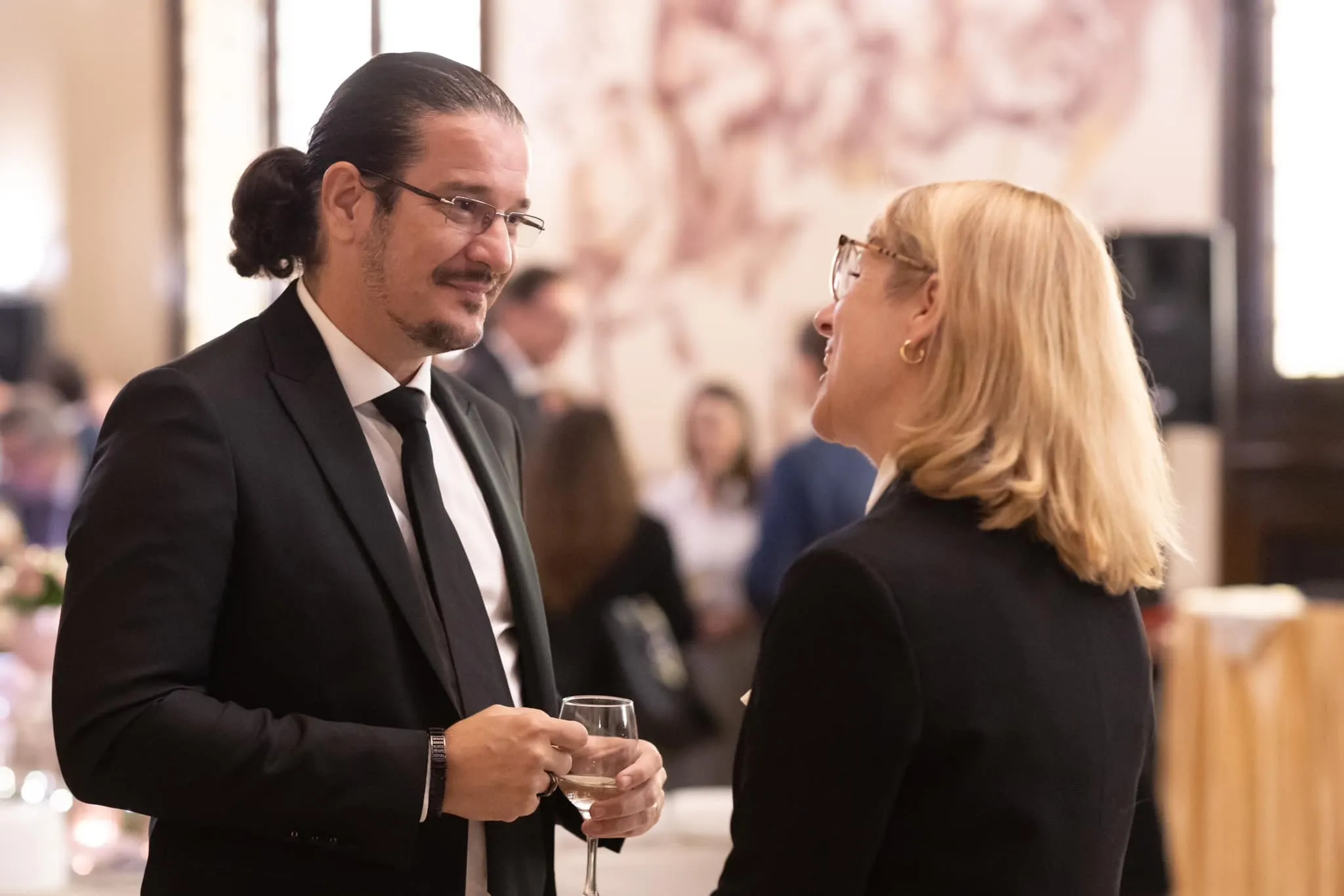 A man in a black suit and a blonde woman in a blazer converse while holding wine glasses at a formal reception.