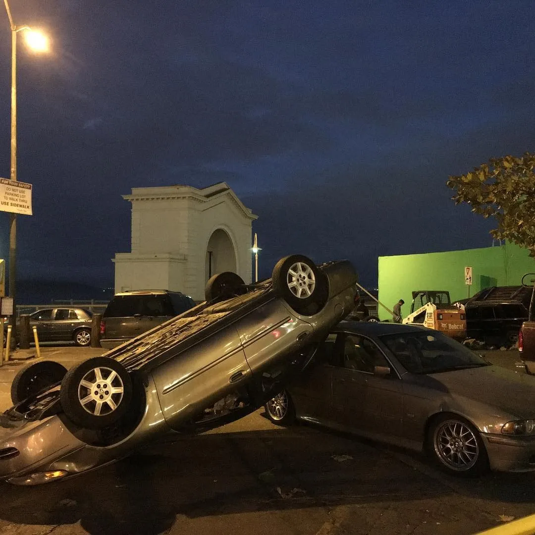 Overturned silver car resting on another car at night beside a white archway and a green screen.
