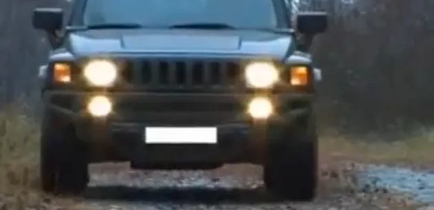 A Hummer with its headlights on approaches on a dirt road.
