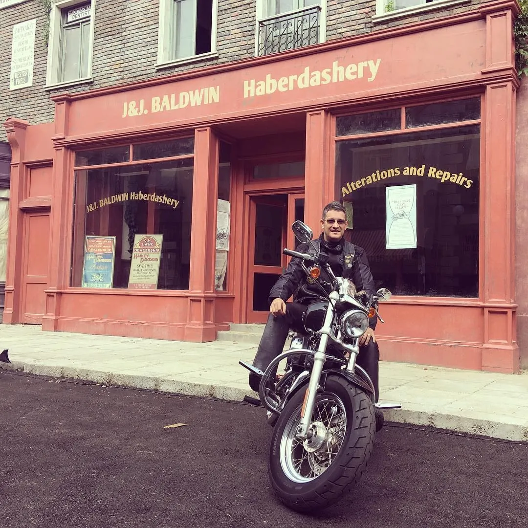 A man in a black leather jacket sits on a motorcycle in front of a red J&amp;J Baldwin Haberdashery shopfront